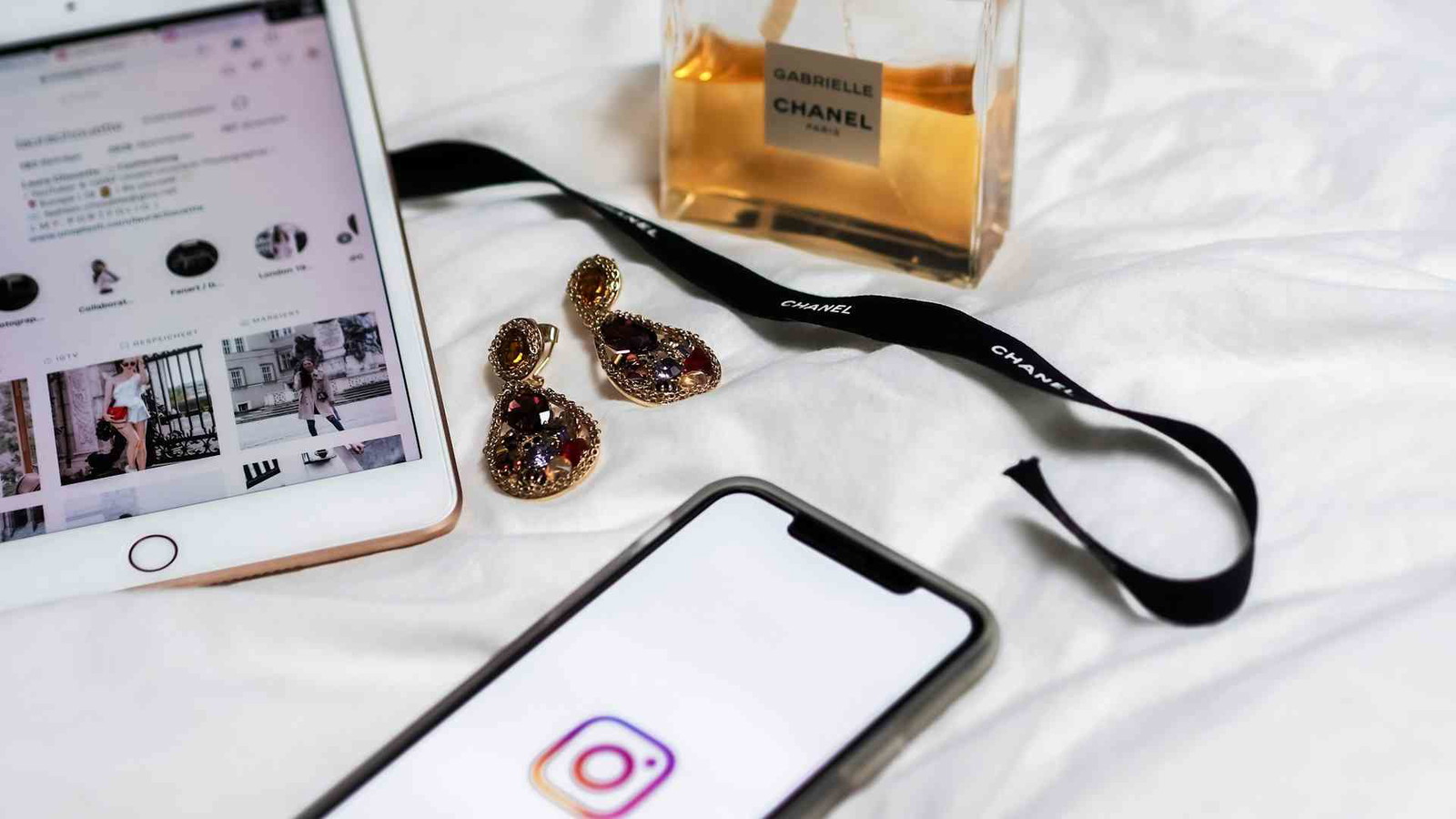 Flat lay of an iPad and smartphone showing Instagram, Chanel perfume, and jewelled earrings on white bedding